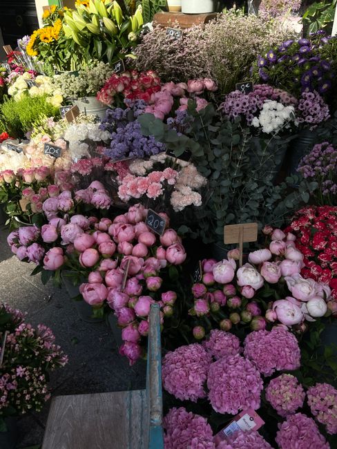 An outdoor display of fresh flowers at a flower market, featuring a variety of blooms in pastel pinks, purples, and whites. There are pink peonies, hydrangeas, and clusters of small purple and lilac b