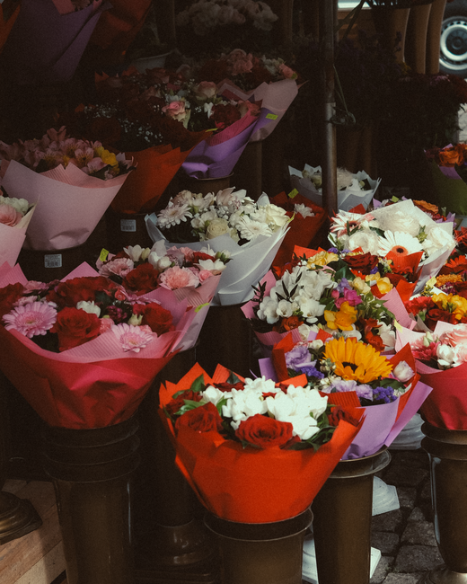 A display of multiple floral bouquets wrapped in colorful paper, featuring a variety of fresh flowers such as roses, gerbera daisies, carnations, and freesias in shades of red, pink, white, yellow, an