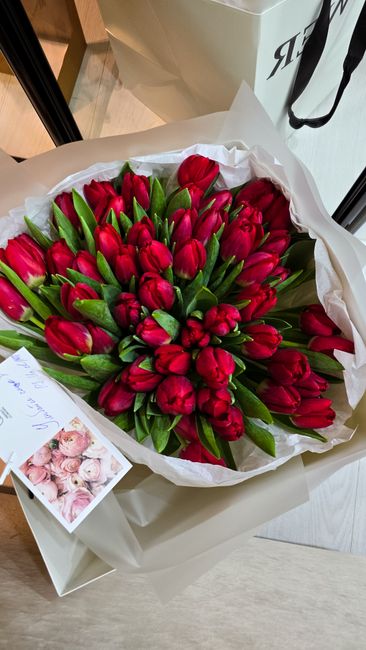 A large bouquet of vibrant red tulips with lush green leaves, arranged in a dense cluster. The flowers are wrapped in white tissue paper with a subtle glossy finish and placed in a white box, suggesti